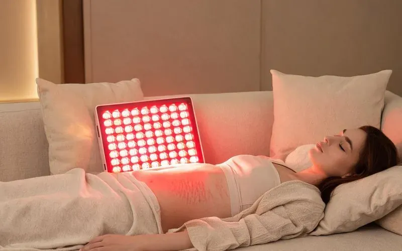 Femme allongée détendue, peau du ventre éclairée par la lueur rouge douce d'un panneau LED, illustration du traitement des cicatrices et vergetures par lumière rouge